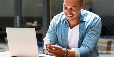 photo of man looking at mobile phone, sitting in front of open laptop