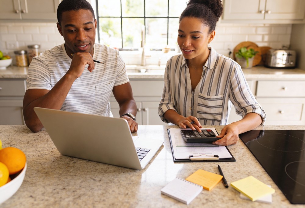 Two people working together at a kitchen island: man using a laptop while woman uses a calculator and takes notes